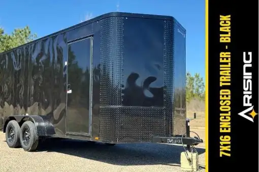Black enclosed trailer with dual axles and perforated steel door parked on gravel with trees in background