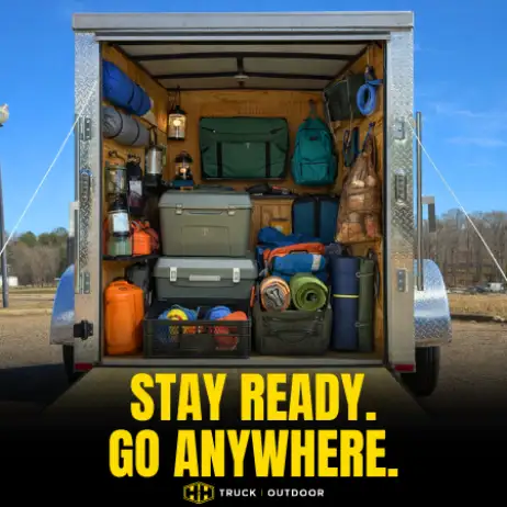 Open trailer packed with camping gear including coolers, tents, sleeping bags, backpacks, and storage containers arranged