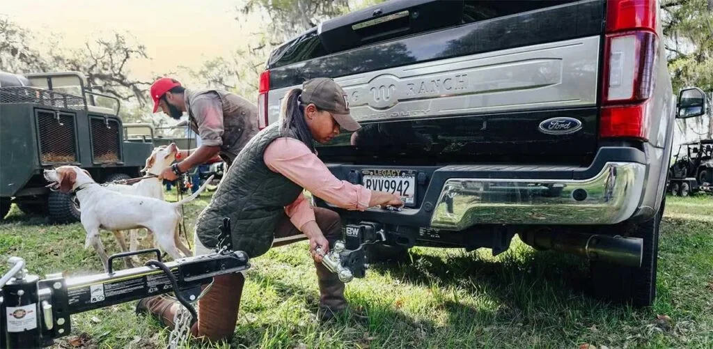 Man crouching beside Ford truck bed, adjusting ball hitch receiver with dog standing nearby on grass