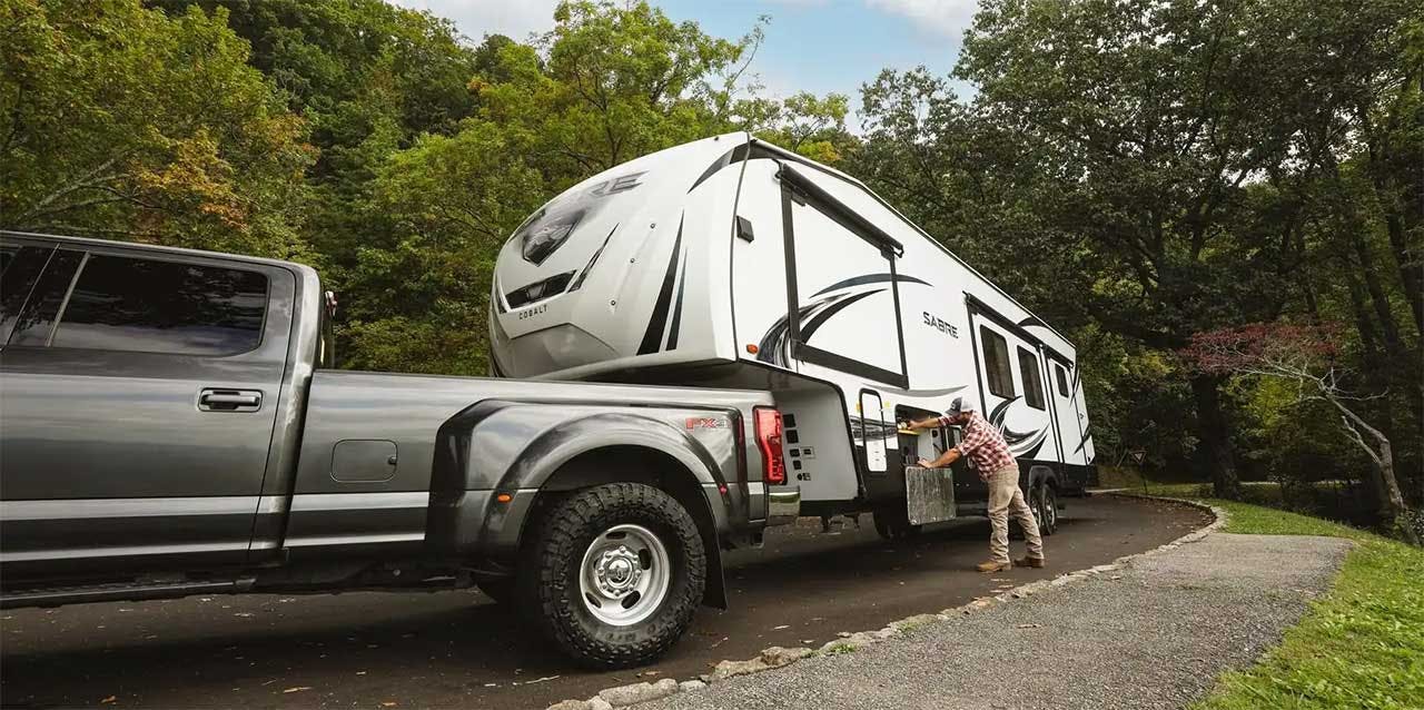 Silver pickup truck towing white travel trailer down a tree-lined driveway