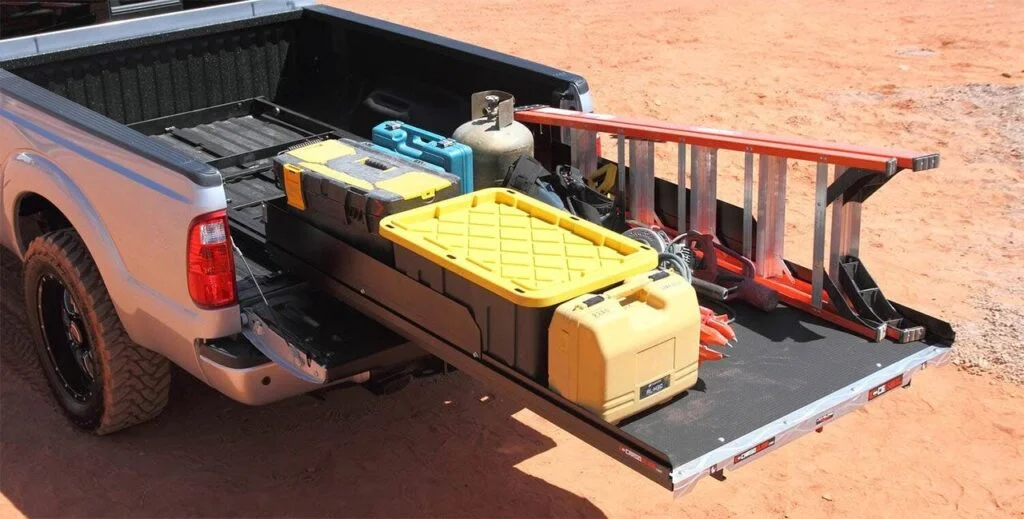 Yellow and black truck bed slide drawer extended from a pickup truck bed, with toolboxes and equipment visible in the truck