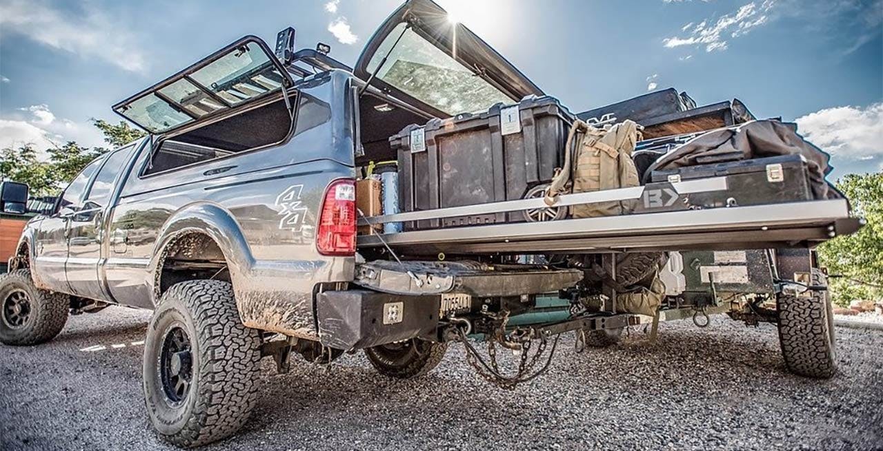 Silver pickup truck with open doors, extended truck bed slide drawer loaded with luggage, toolboxes, and gear in desert