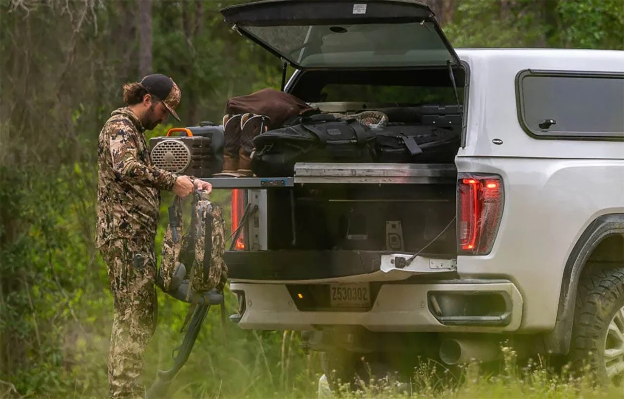 Person loading a dark slide-out bed tray mounted on white pickup truck bed near a forest setting