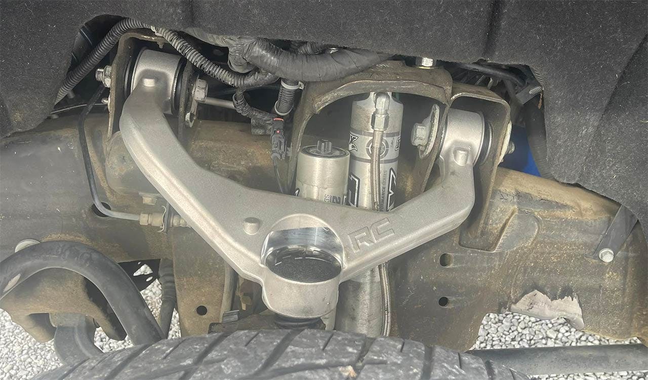 Underside view of truck suspension showing control arms, ball joints, and connected hardware with visible rust and wear.