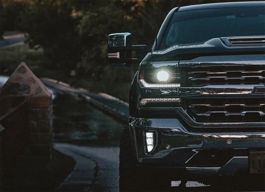 Black pickup truck with headlights on, parked near evergreen trees and a cabin in the background