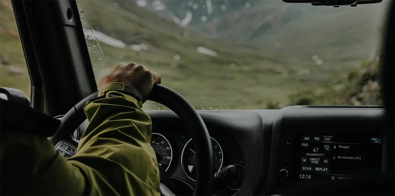 Driver in olive jacket grips steering wheel inside truck cab during daytime drive through mountainous terrain