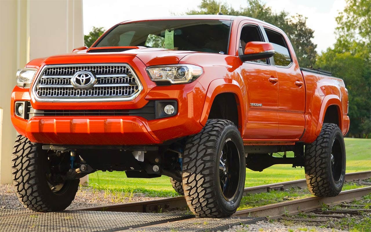 Orange Toyota Tacoma truck lifted on oversized black all-terrain tires in a residential driveway with trees in background.