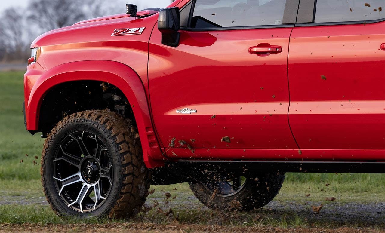 Red lifted pickup truck with oversized all-terrain tires and custom black wheels parked on grass near a road