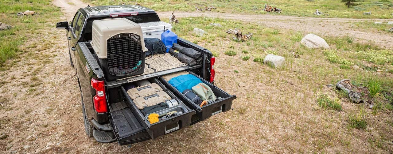 Truck bed with dual sliding drawers open, showing pet carrier, tools, and gear stored inside, parked at outdoor campground