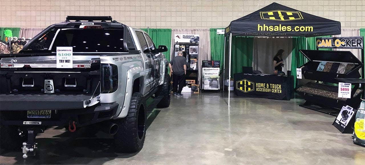 Black pickup truck with roof rack displayed next to Home & Truck Accessory Center vendor booth at indoor exhibition.