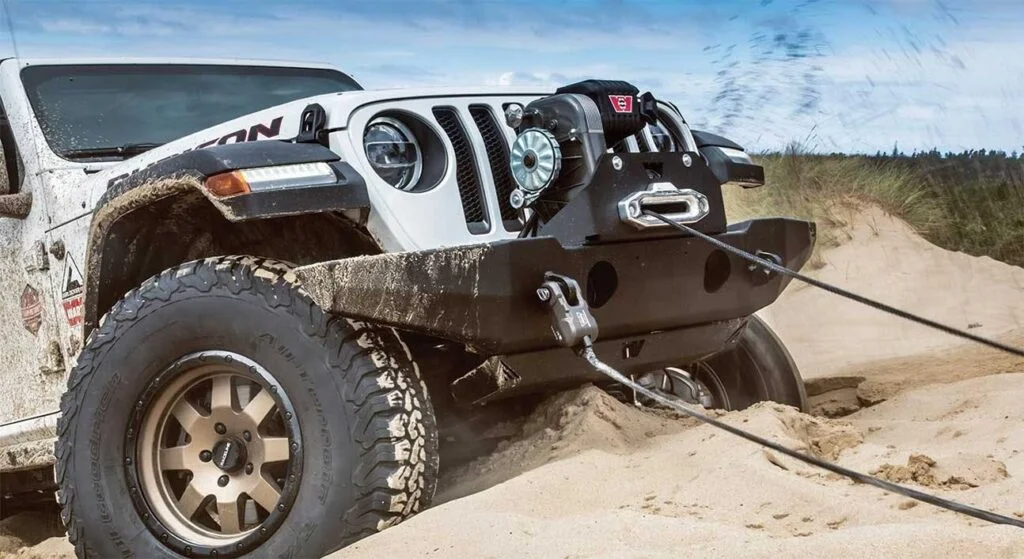 White Jeep stuck in sand with black winch mounted on front bumper, cable extended to anchor point in sandy terrain