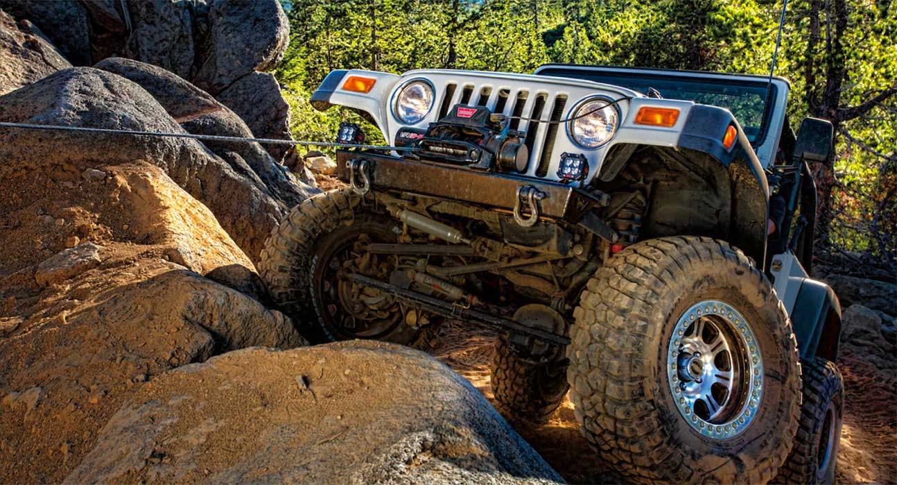 Lifted Jeep with winch mounted on front bumper navigating between large boulders on rocky forest trail.