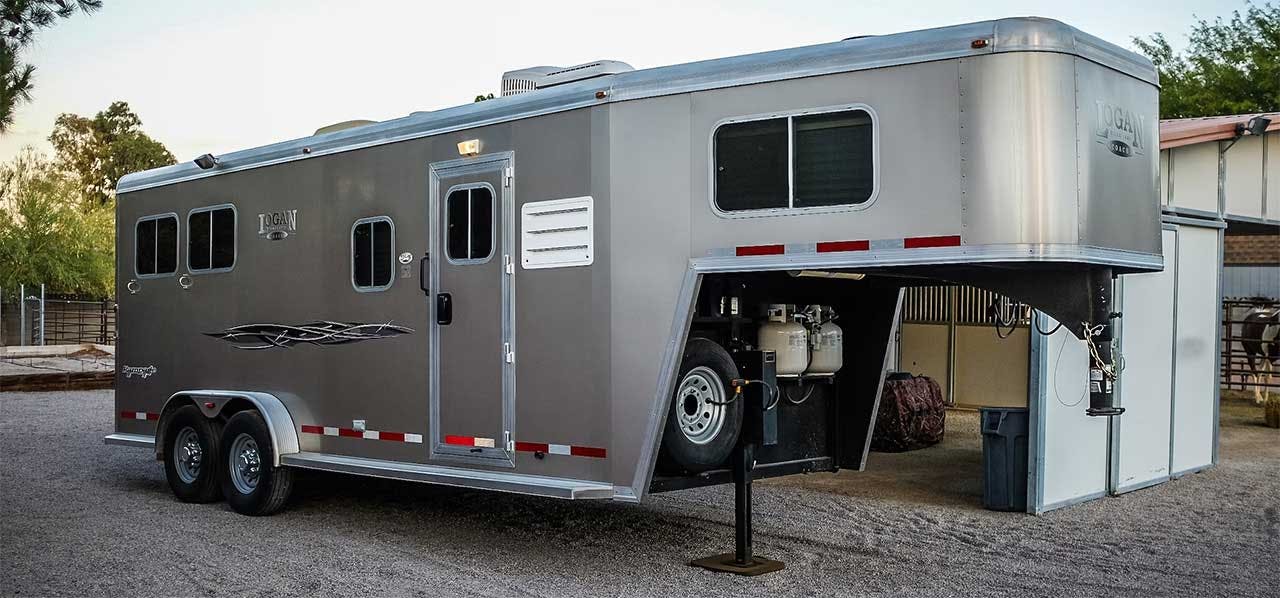 Silver livestock trailer with open side compartment parked at a rural facility with fencing and buildings visible in
