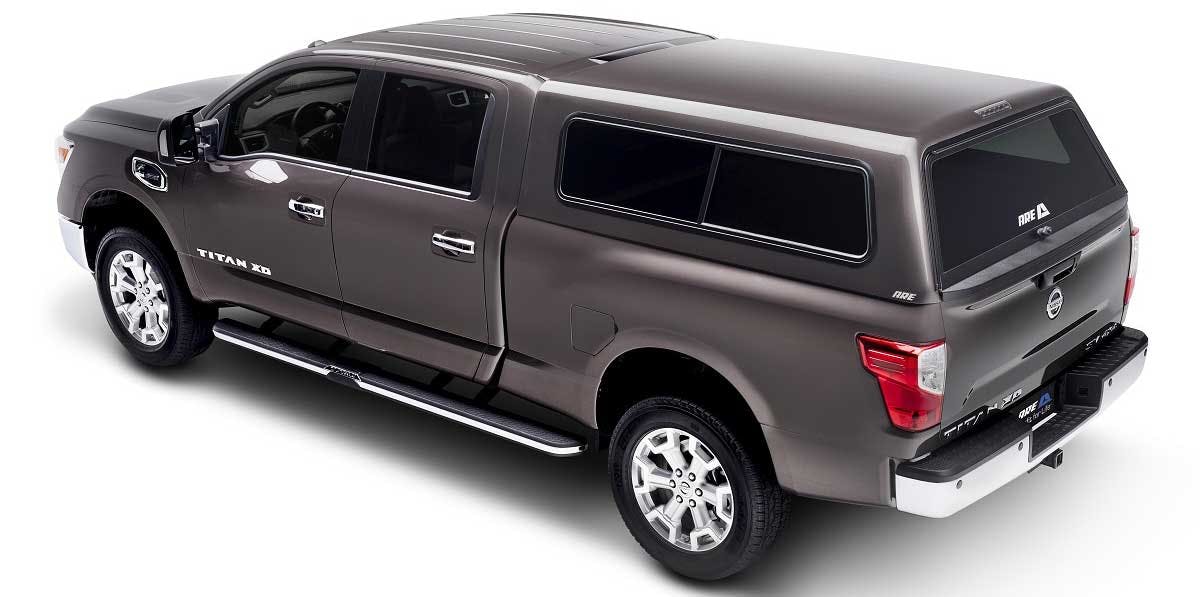 Dark gray Nissan Titan XD crew cab with open driver door and black A.R.E. camper shell with roof rack installed on bed