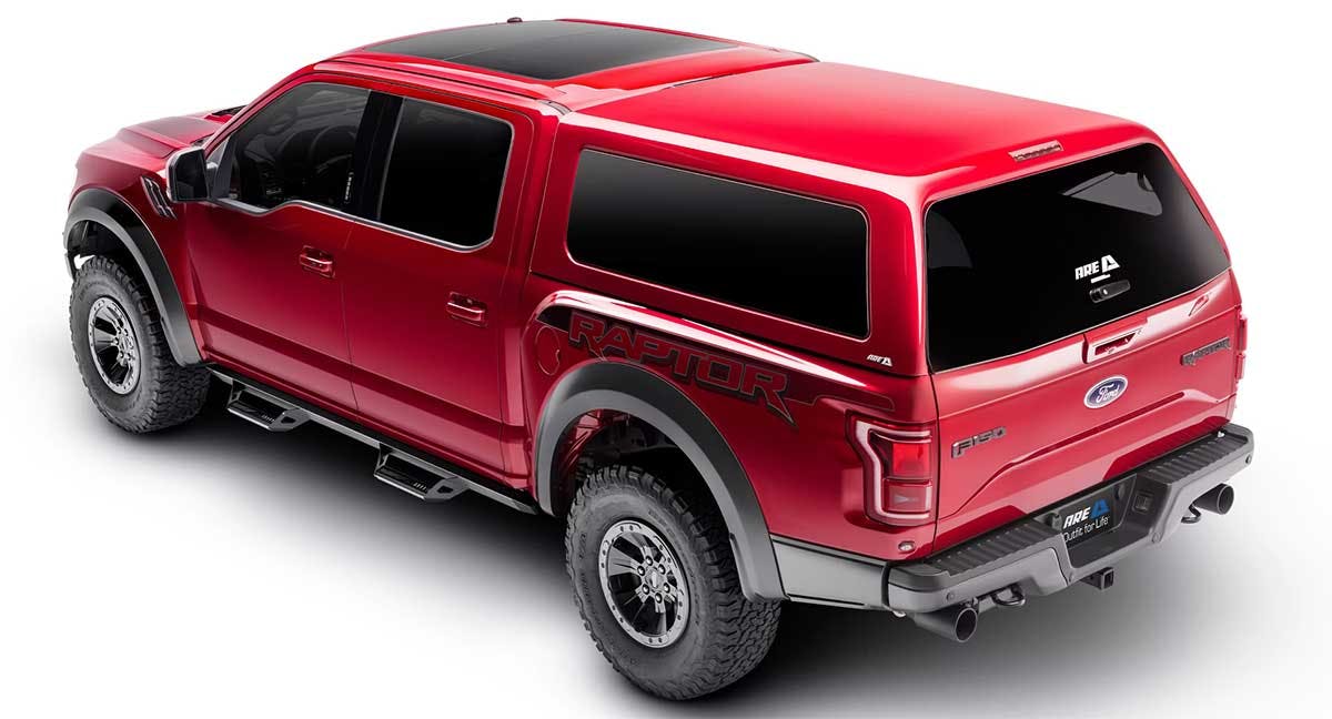 Red Ford F-150 Raptor pickup truck with A.R.E. camper shell mounted on the bed, viewed from rear three-quarter angle.
