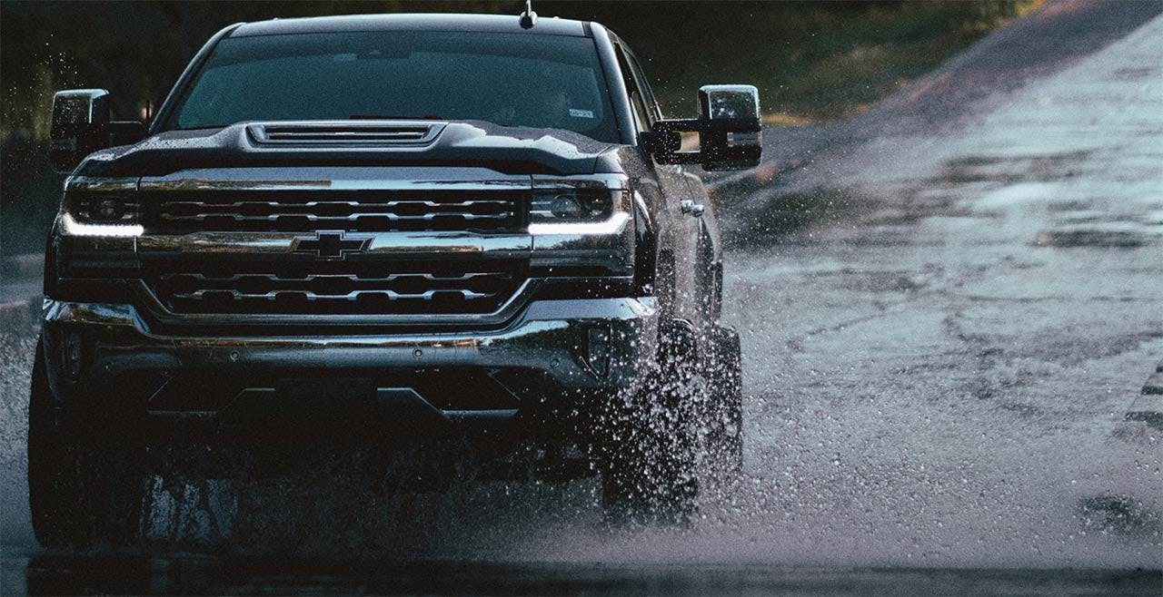 Black Chevrolet truck driving through water on dirt road, splashing muddy water from tires and undercarriage.