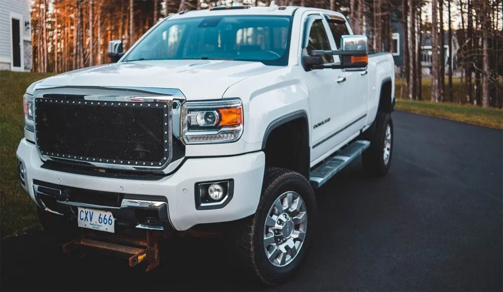 White GMC Sierra truck with black mesh grille and chrome accents parked on driveway with pine trees in background