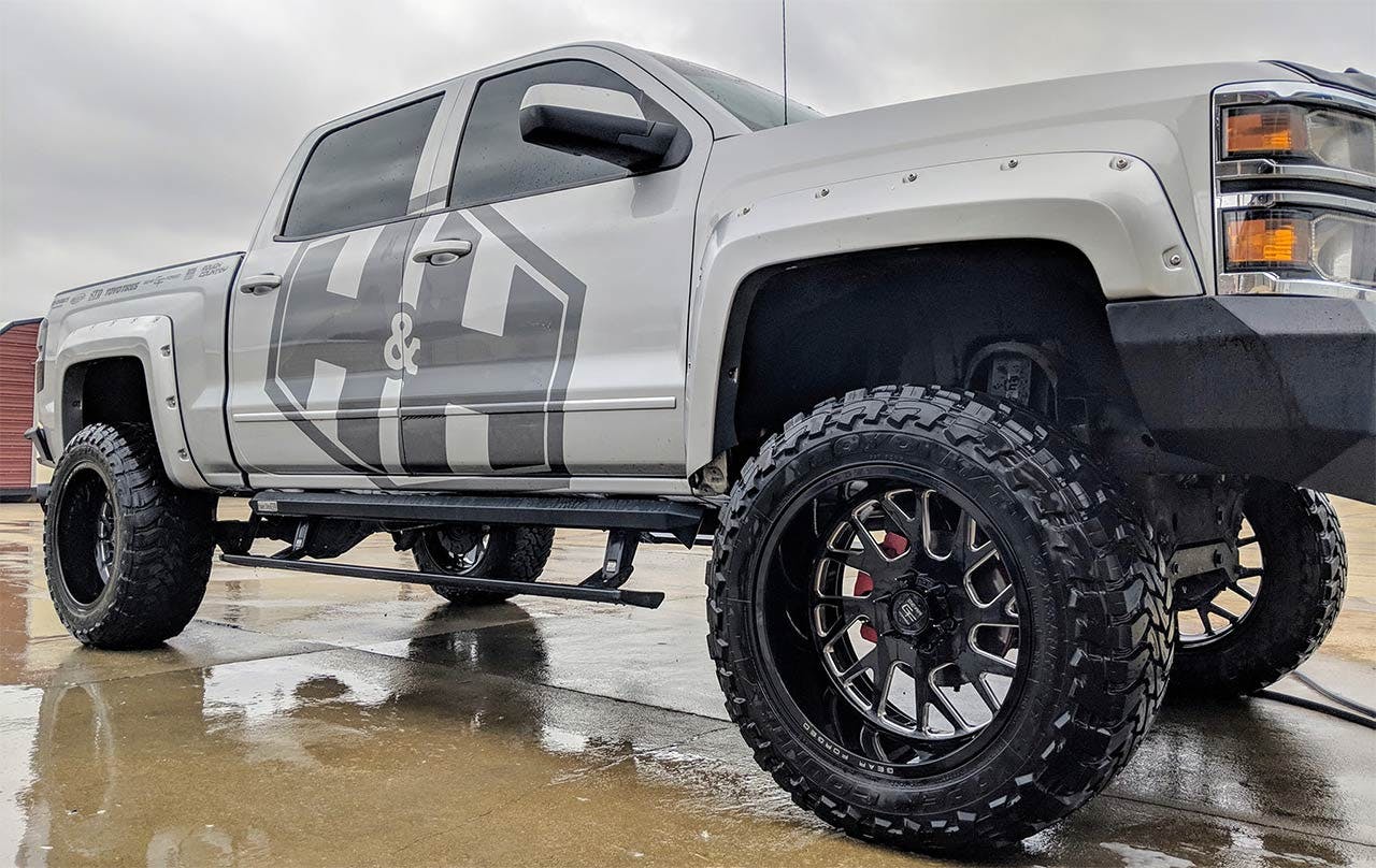 White lifted pickup truck with custom striped graphics parked on wet pavement, showing oversized all-terrain tires on black