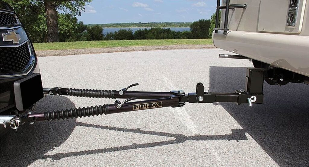 Blue Ox tow bar attached between a silver RV and tow vehicle at a lakeside rest stop with trees and water visible behind