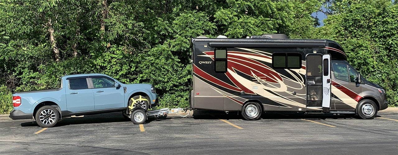 Light blue pickup truck parked next to a burgundy and cream motorhome in a parking lot surrounded by trees.