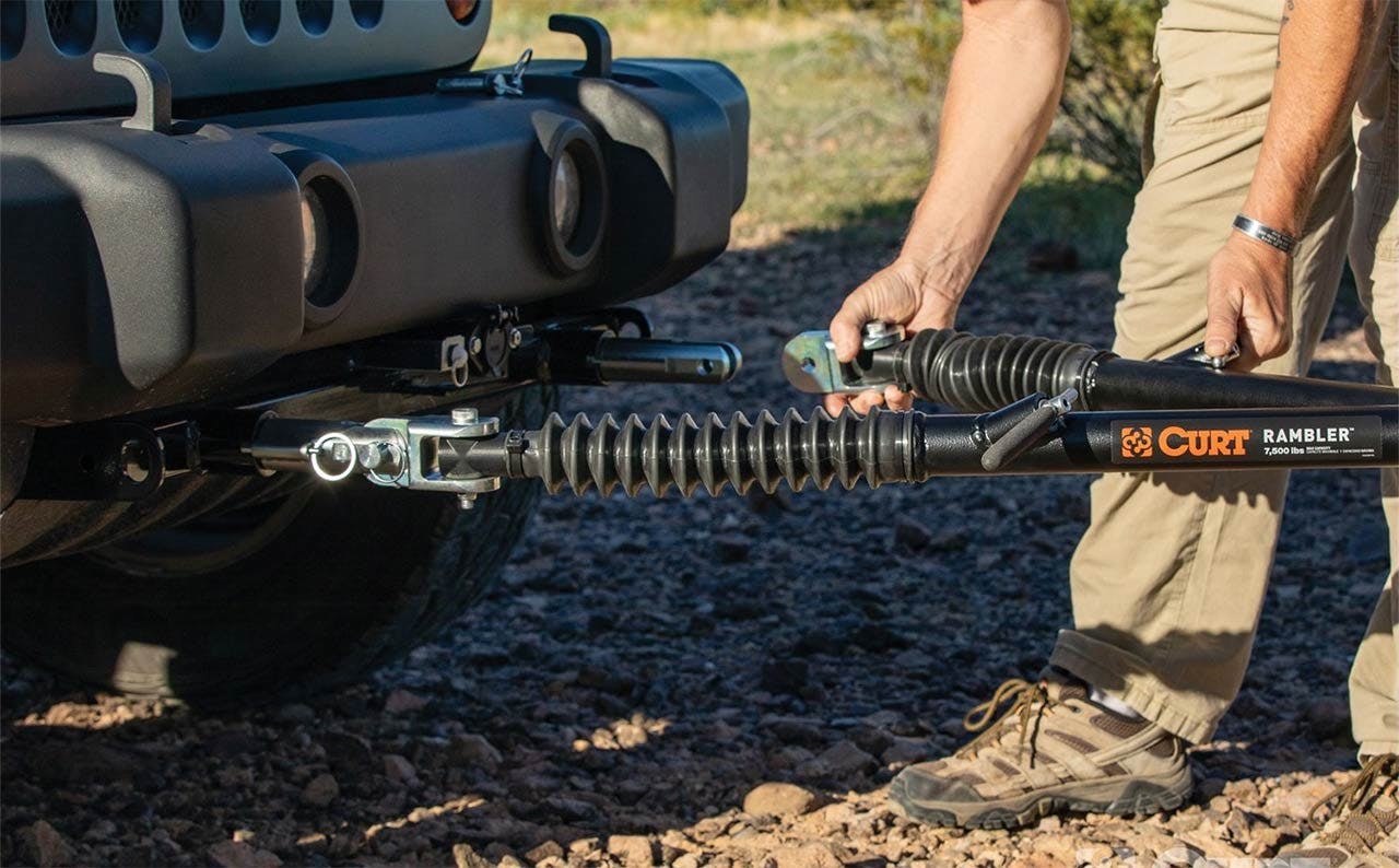 Person holding a CURT Rambler tow bar with coiled spring suspension connected to a truck hitch on rocky ground.
