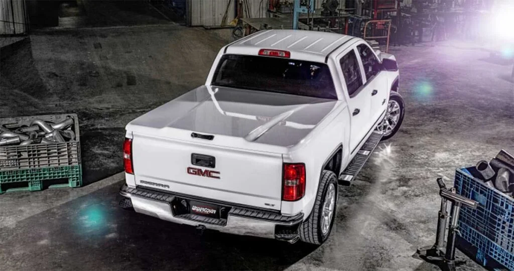 White GMC pickup truck with an open hard tonneau cover revealing the empty bed in an industrial garage setting