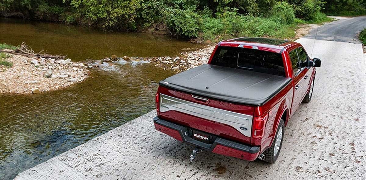 Red pickup truck with gray tonneau cover parked on gravel beside a river with dense green trees lining the bank.