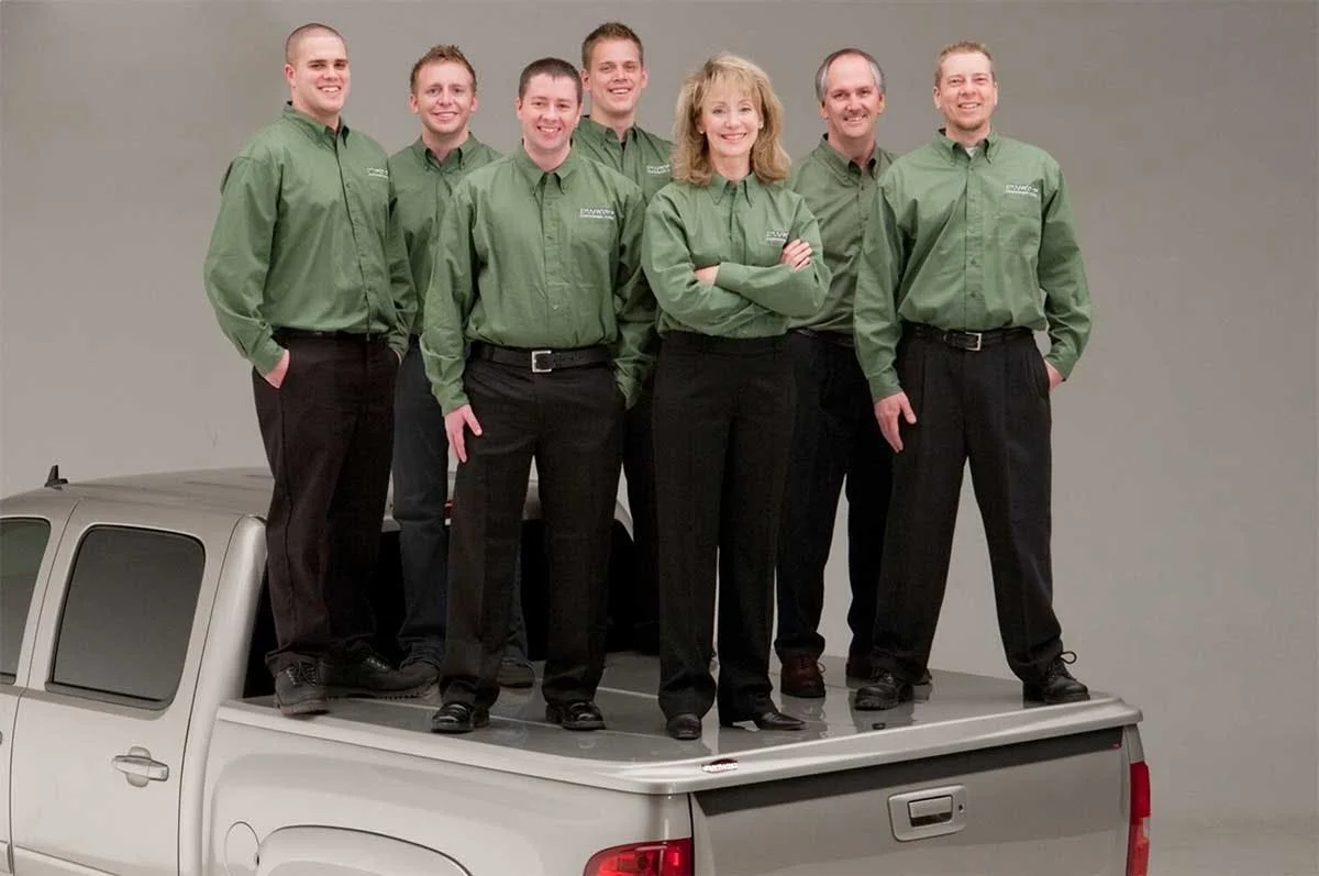 Seven employees in matching green work shirts standing on the bed of a white pickup truck.