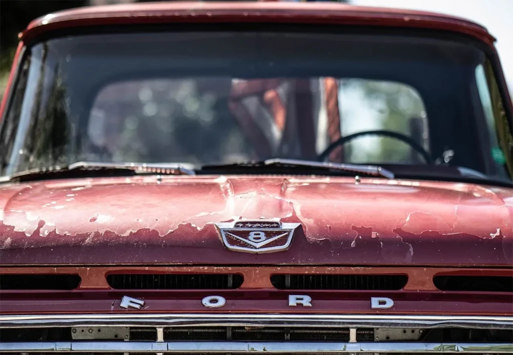 Faded red paint on vintage Ford truck hood with peeling clear coat and V-8 emblem visible