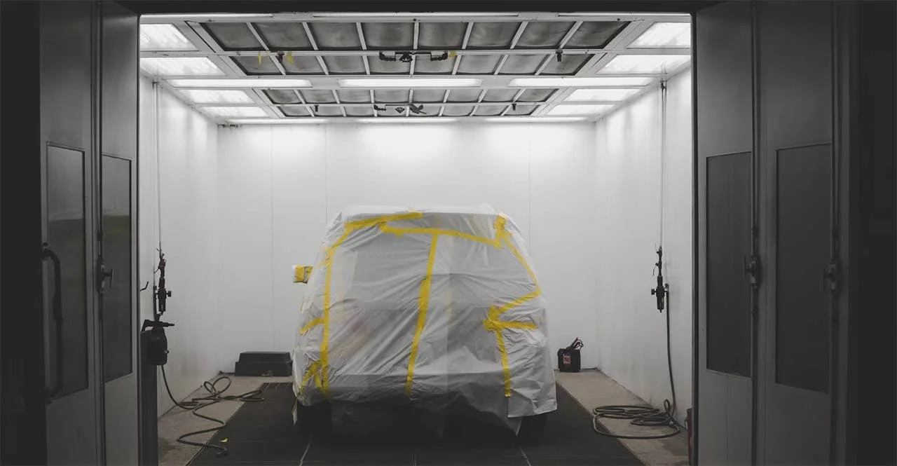 Truck covered in plastic sheeting with yellow tape straps in a professional paint booth with fluorescent lighting overhead.