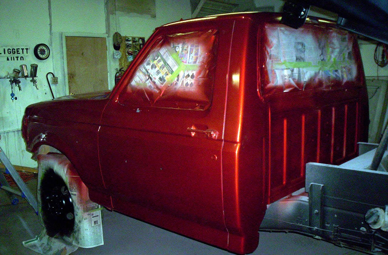 Red truck cab in a garage workshop with windows showing preparation notes, surrounded by work tools and equipment on walls