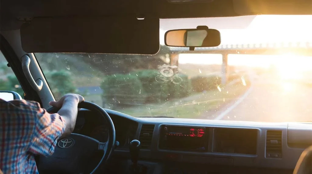 Driver in plaid shirt at steering wheel with bug-spattered windshield during sunset drive