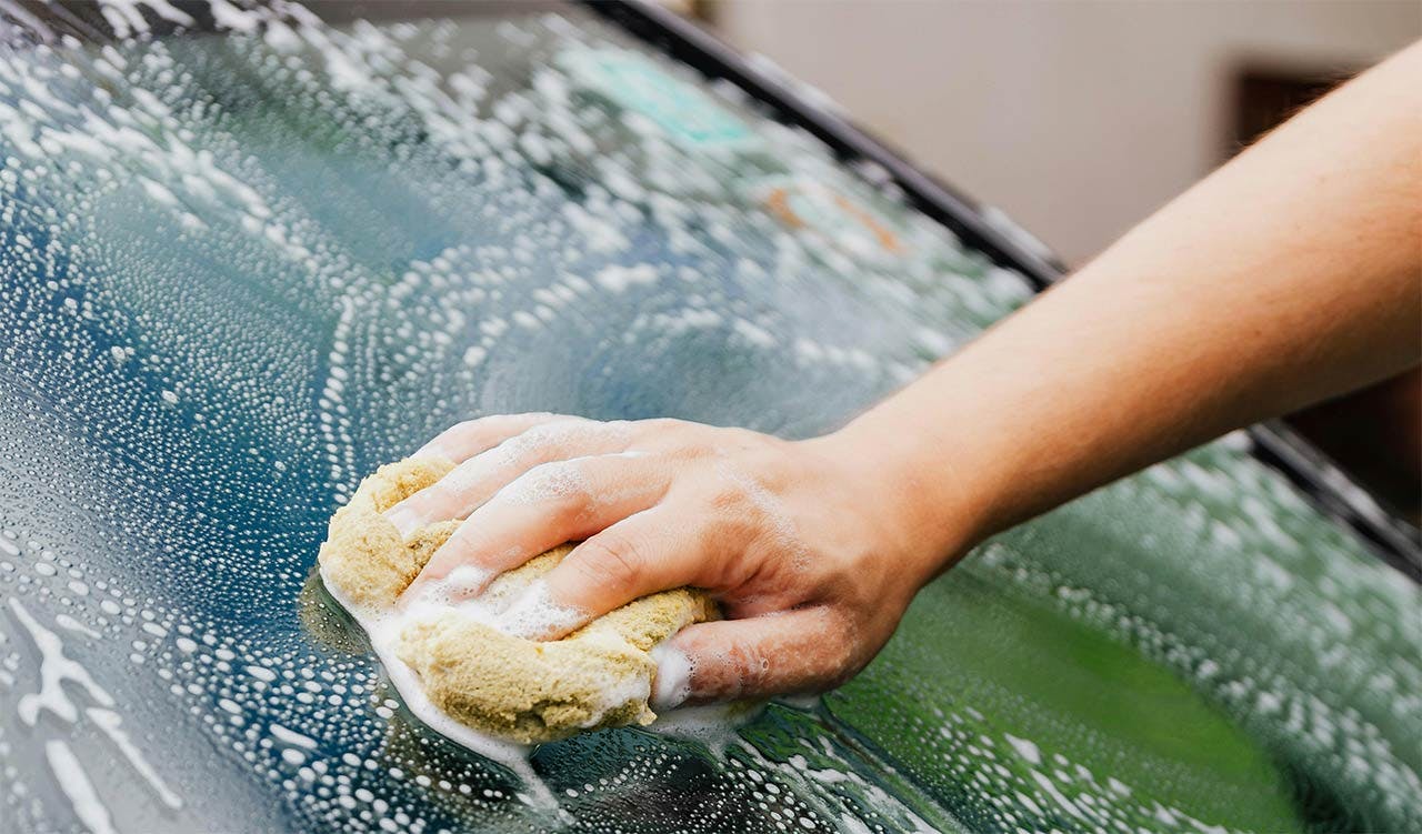 Hand holding a wet soapy sponge scrubbing a wet truck hood covered in water droplets.