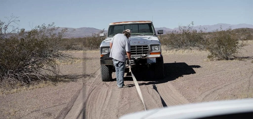 Person standing beside a truck on a desert road, connecting a towing cable to the front bumper.