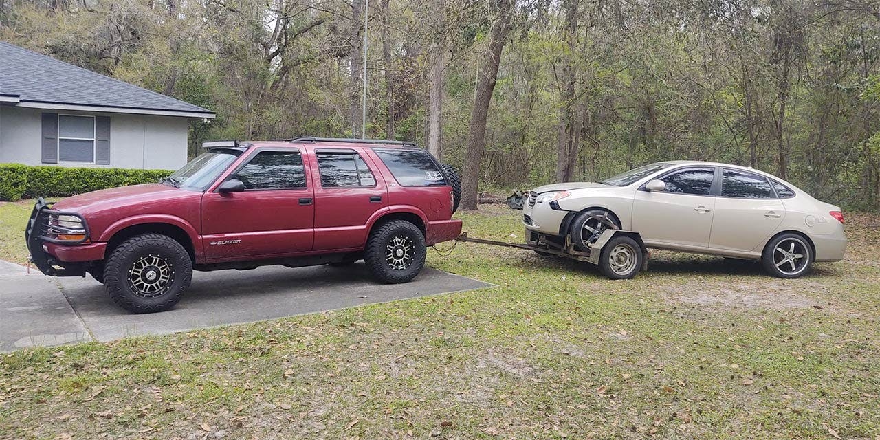 Maroon lifted truck connected by tow strap to beige sedan in driveway with wooded background