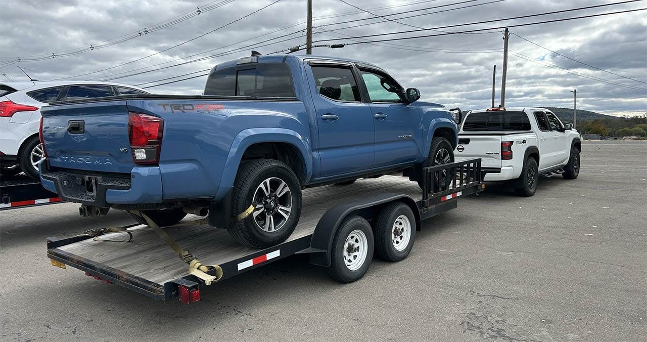 Blue Toyota Tacoma loaded onto a dual-axle flatbed trailer with yellow straps securing the wheels.