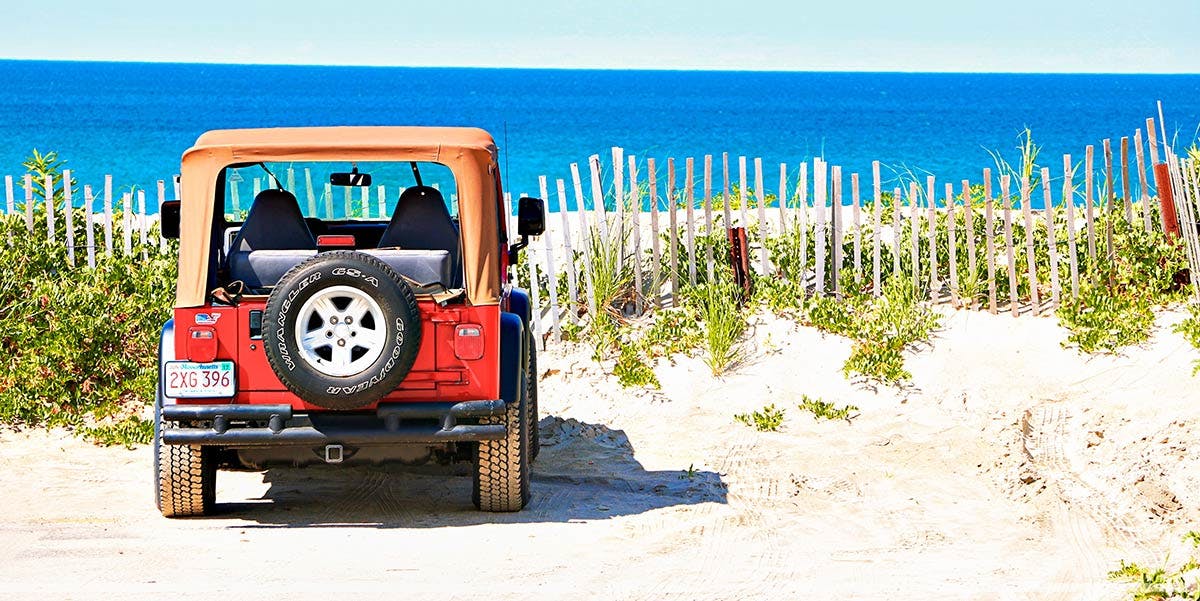 Red Jeep with tan soft top parked on sandy beach with white dune fence and ocean in background