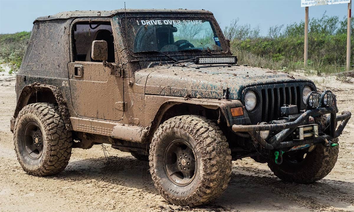 Mud-caked Jeep Wrangler parked on sandy ground with large terrain tires and mounted lights on front bumper