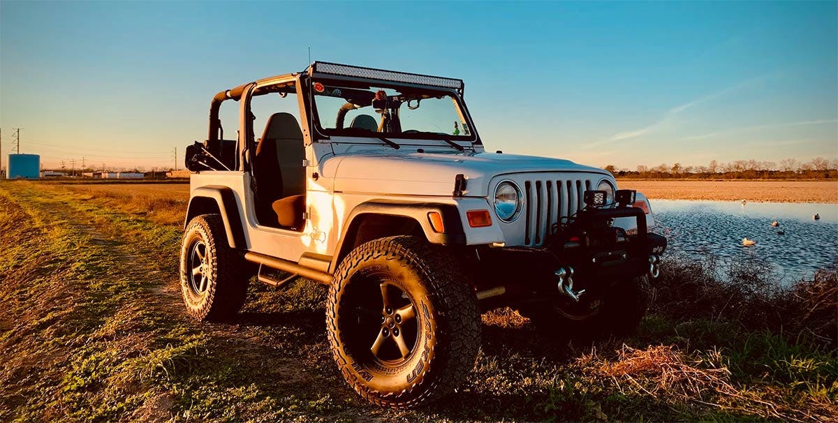 Tan Jeep with doors removed and soft top folded back, parked beside water in an open field at sunset.