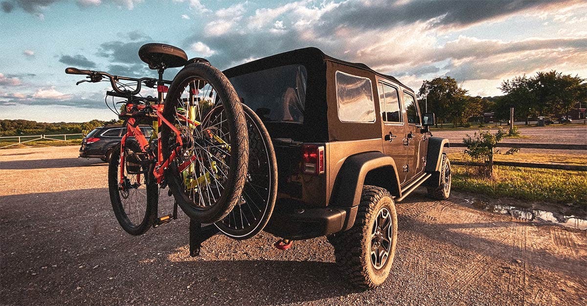 Red bicycle mounted on black rear hitch carrier attached to tan Jeep parked at ranch with open fields and trees.