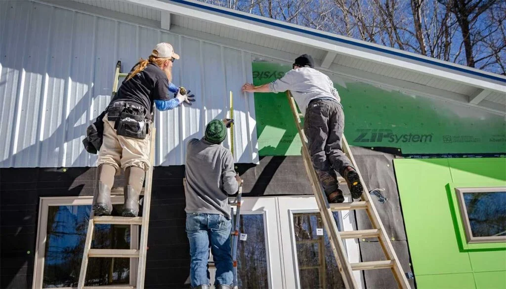 Three workers on ladders installing green metal panels on a white building with a blue trim.