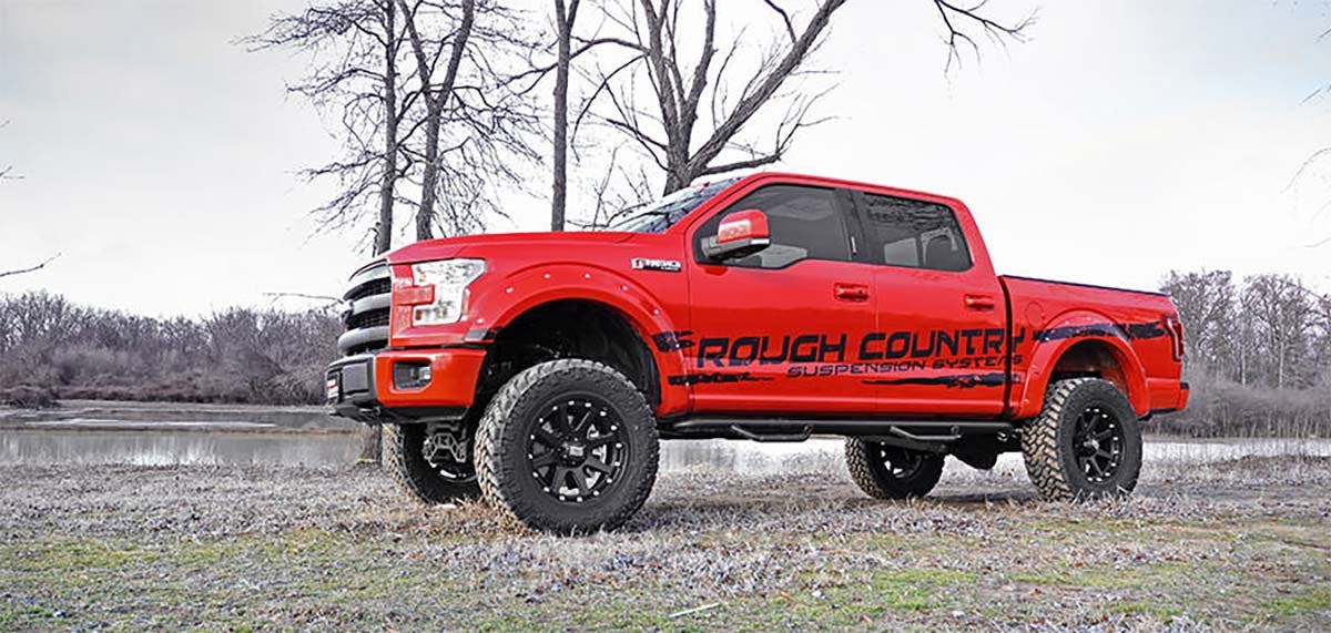 Red lifted pickup truck with oversized black wheels parked on gravel near bare trees and a lake shoreline.