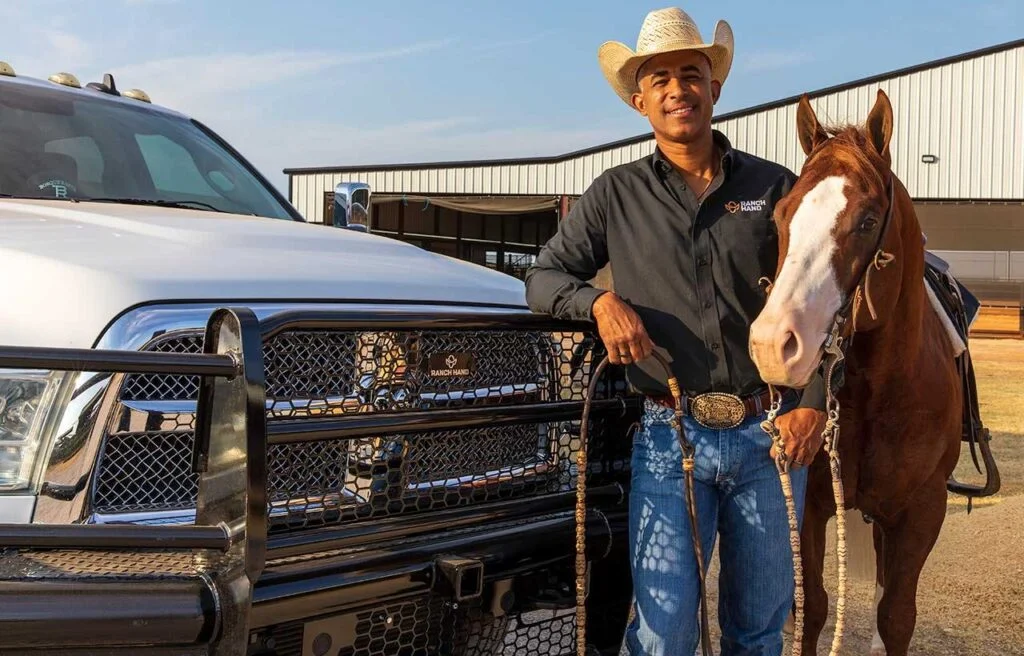Man in cowboy hat standing next to brown horse beside white truck equipped with black metal grille guard and bumper