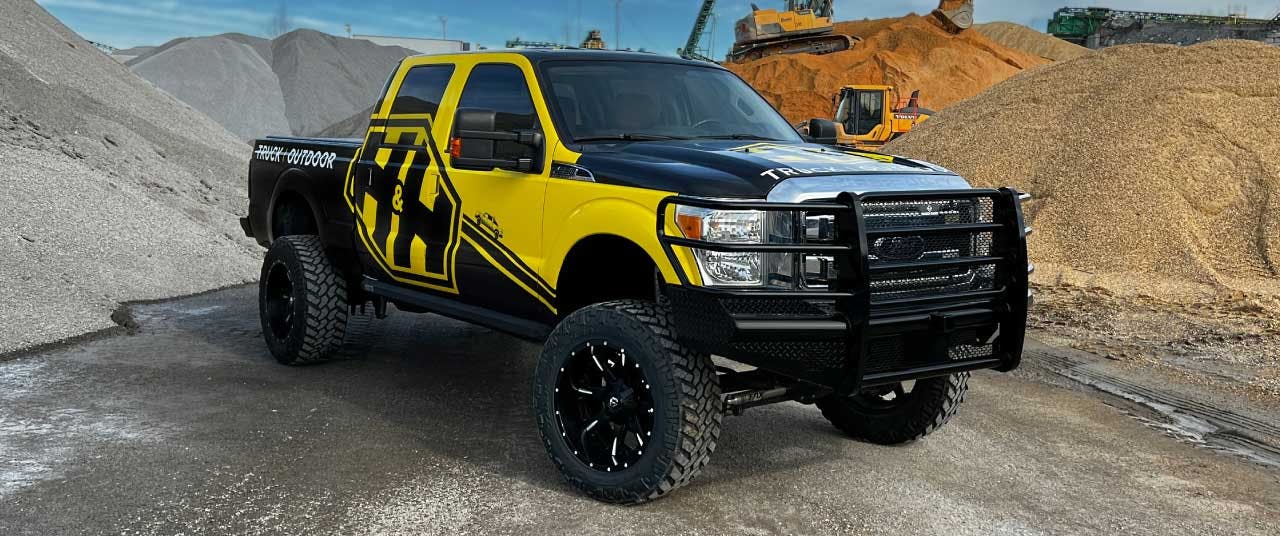 Yellow and black lifted truck with grille guard parked at construction site with aggregate piles and equipment behind it.