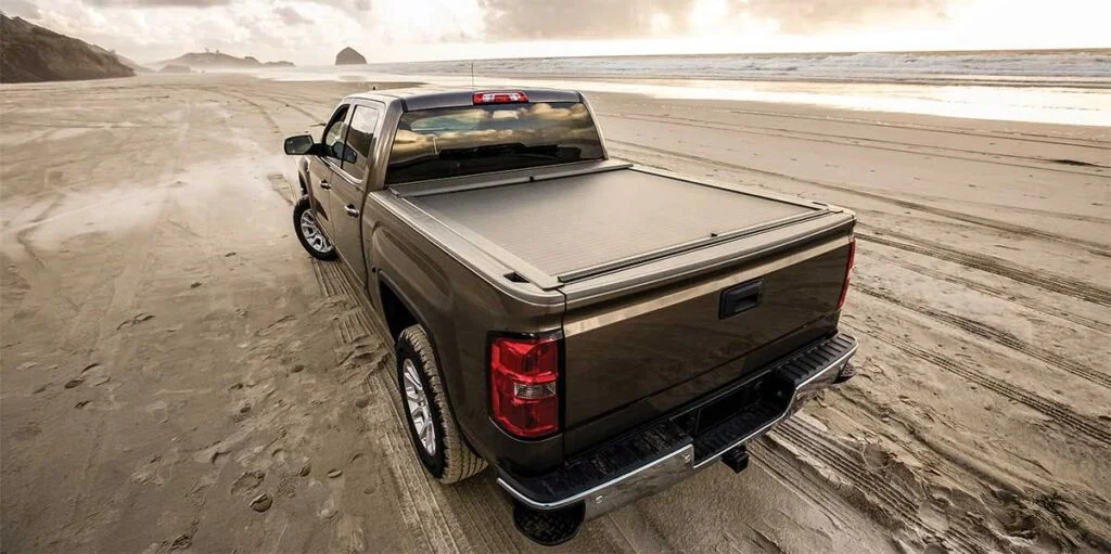 Gray pickup truck with retractable bed cover closed, parked on a wide sandy beach with ocean and cliffs in background.