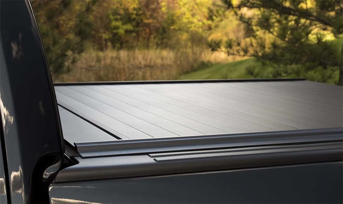 Retractable metal truck bed cover with horizontal slats partially open over truck bed, trees visible in background