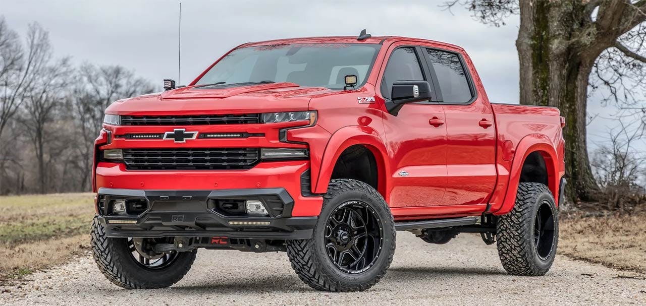 Lifted red Chevrolet pickup truck with black grille and custom wheels on gravel driveway beside bare trees.