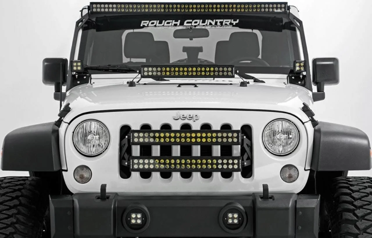 White Jeep with multiple yellow LED light bars mounted on roof, hood, and front bumper grille.
