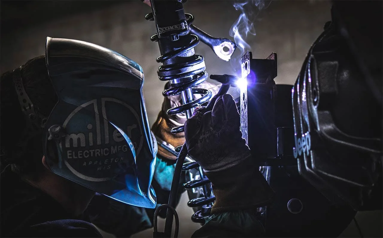 Coilover suspension strut assembly with blue coils and chrome components, welding spark visible on mounting bracket