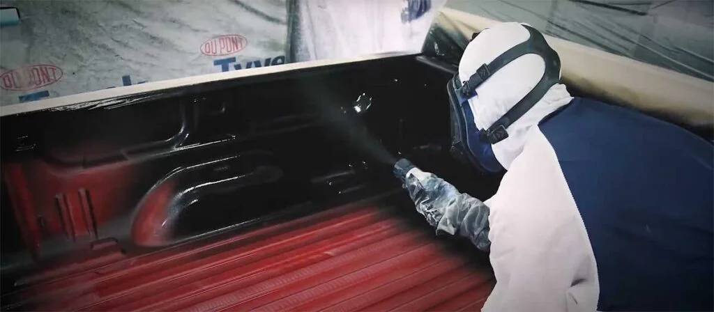 Worker in protective gear spray-applying black coating to inside of red truck bed